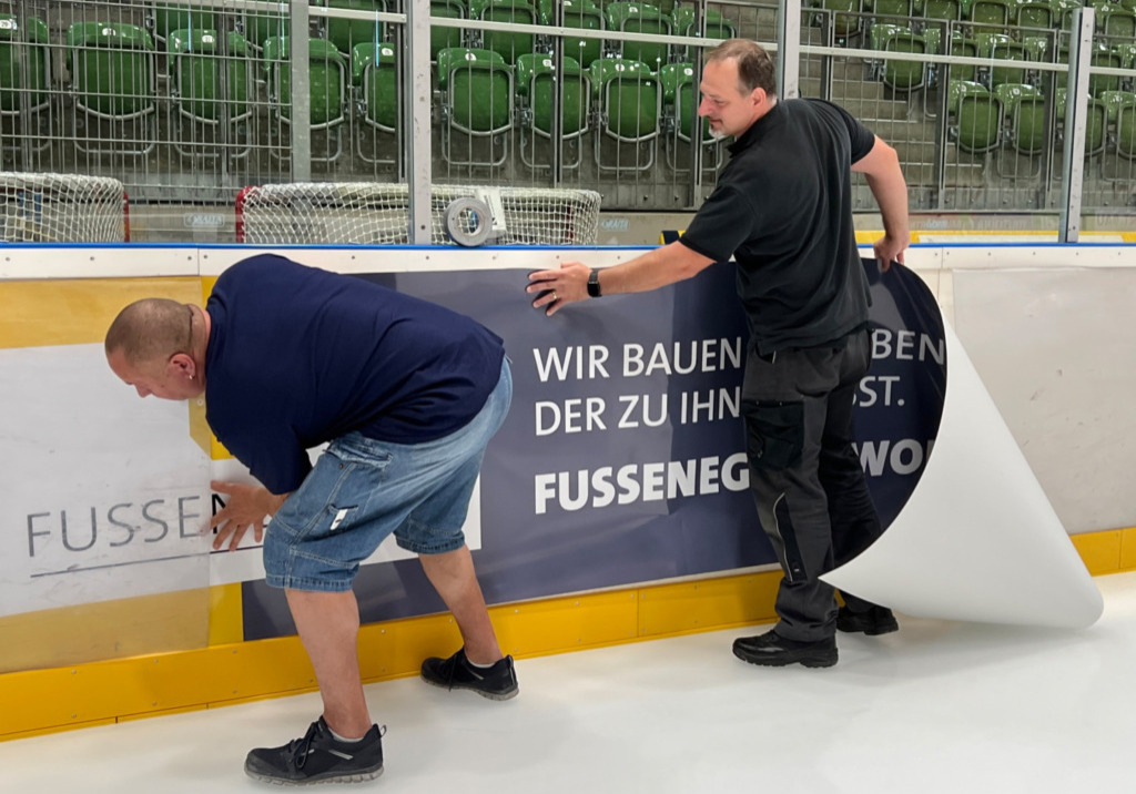 Mit Hilfe der beiden Eismeister zog die Fussenegger Wohnbau GmbH über die neue Bandenwerbung in die CHG Arena ein. Bild: Fussenegger Wohnbau GmbH